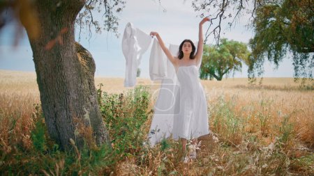 Gorgeous model posing country exterior. Confident young woman raising hands standing near white linen on rope at spikelets field. Seductive girl turning back touching clean drying laundry at meadow