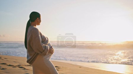 Schwangere bei Sonnenuntergang am schönen Sandstrand. Friedliche werdende Mutter, die vor Meereswellen steht und den Sonnenuntergang betrachtet. Ruhiges Mädchen streichelt Bauch und genießt Schwangerschaft an der Küste