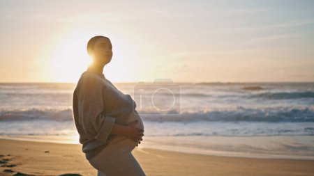 Werdende Mutter posiert bei Sonnenuntergang am Sandstrand und streichelt Bauch. Ruhige inspirierte Frau streichelt schwangeren Bauch vor schönen Meereswellen am Abend. Mädchen erwartet Baby genießen Sonnenuntergang in der Natur.