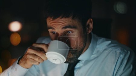 Travailleur des heures supplémentaires boire du café assis bureau sombre seul fermer. Portrait d'homme barbu bourreau de travail travaillant sur ordinateur portable tard dans la soirée. Entrepreneur concentré occupé se dépêchant d'accomplir la tâche de travail la nuit.