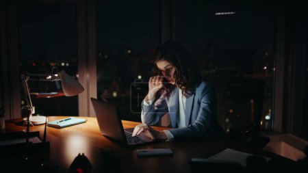 Sleepy businesswoman working at night sitting office desk. Young woman manager feeling tired looking laptop monitor. Upset lady employee overwhelmed by overwork late evening. Busy company worker.