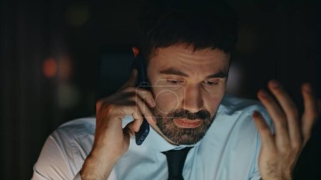 Exhausted employee talking smartphone solving business problem at night close up. Tired bearded manager calling worried work troubles sitting office late evening. Frustrated worker man speaking phone.