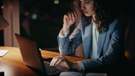 Business woman busy of overtime work sitting dark office workplace close up. Attractive young girl manager typing report on laptop feeling tired. Serious elegant businesswoman overworking at night.