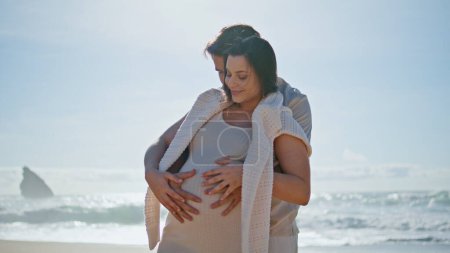 Paar erwartet Baby am sonnigen Strand und streichelt dicken Bauch zusammen. Glückliche zukünftige Eltern genießen das Familienwochenende am Sommerstrand. Liebender zarter Mann umarmt geliebte schwangere Frau