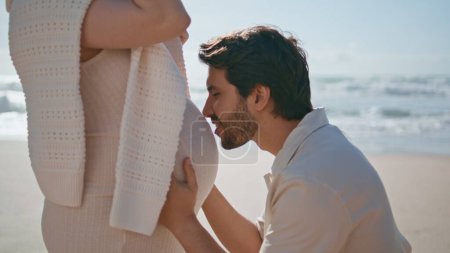 Husband kissing pregnant belly wife standing on sunny seashore close up. Loving man closing eyes bonding to big tummy on sandy beach. Future father expressing love to expected baby. Pregnancy concept