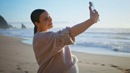Pregnant woman making selfie standing beautiful sandy seashore caressing belly close up. Carefree happy girl expecting baby taking photography big tummy using smartphone. Smiling lady enjoy pregnancy.
