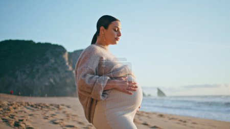 Woman touching pregnant belly standing on picturesque shoreline sunny evening close up. Happy carefree expectant mother posing on sand beach caressing tummy. Smiling tender lady enjoy pregnancy.