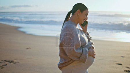 Zukünftige Mutter spricht Smartphone zu Fuß schönen Strand aus nächster Nähe. Lächelnde, glückliche Schwangere, die mit dem Handy am goldenen Sandstrand anruft. Fröhliche Dame erwartet Baby und streichelt Bauch.