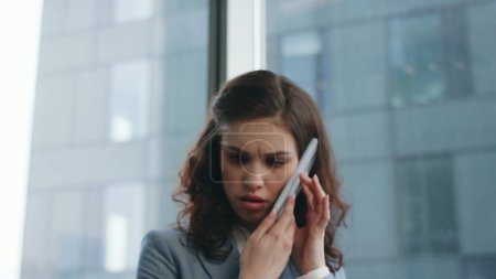 Stressed businesswoman speaking phone checking work papers on desk close up. Worried business lady talking smartphone at modern workplace feeling irritated. Nervous woman confused by telephone call.