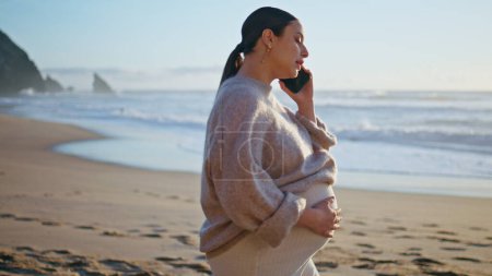 Pregnant woman calling cellphone at beach caressing big belly. Happy carefree girl expecting baby talking smartphone walking sand coast. Smiling future mother enjoy phone communication at nature