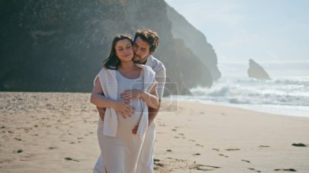 Hugging pregnant couple walking beach in front beautiful ocean waves. Happy affectionate spouses waiting baby relaxing together at sunny seashore. Handsome husband embracing wife holding big belly.