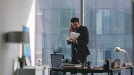 Excited broker laughing calling phone standing modern office. Emotional businessman throwing papers up rejoicing great deal sharing good news by telephone. Happy ceo feeling success talking cellphone.