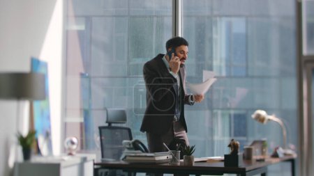 Businessman talking cellphone nervously standing office looking documents. Angry bearded manager shouting on phone conversation at modern workplace. Man entrepreneur call dissatisfied business report.