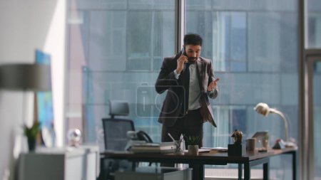Employee frustrated by phone call standing office looking business documents. Angry bearded businessman arguing on telephone conversation at workplace. Stressed manager speaking smartphone nervously.