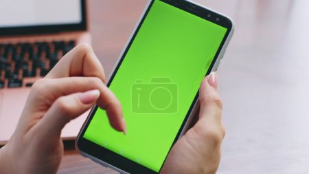 Unrecognizable woman using smartphone with green screen on modern workplace close up. Girl hands scrolling tapping on chroma key telephone. Unknown lady looking mobile banking application at home.