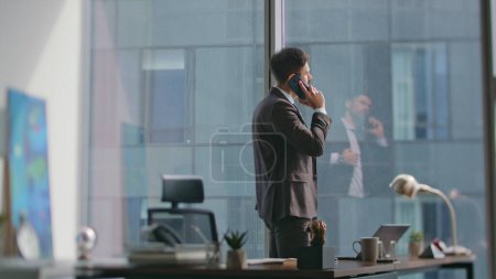 Elegant business man calling partners standing luxury office sunny morning. Confident bearded man entrepreneur talking cell phone writing notes at work desk. Busy ceo making appointment by telephone.