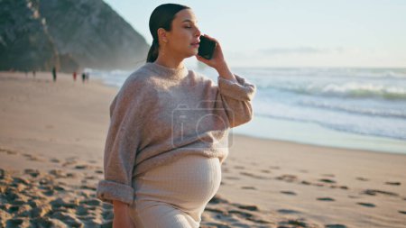 Schwangere, die am Strand anruft und ihren dicken Bauch streichelt. Glückliche unbeschwerte Frau erwartet Baby reden auf dem Smartphone zu Fuß schönen Sandküste. Zarte zukünftige Mutter lächelt und genießt die Telefonkommunikation.