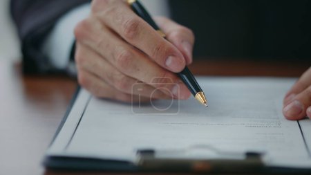 Businessman hand holding pen signing report after checking information close up. Unknown manager reading contract terms closely putting signature on paper. Office worker certifying business deal.
