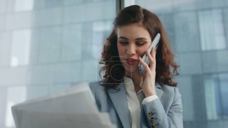 Office manager talking cellphone with smile looking at business documents close up. Portrait attractive happy woman consultant speaking phone in modern office window. Cheerful girl enjoy conversation.