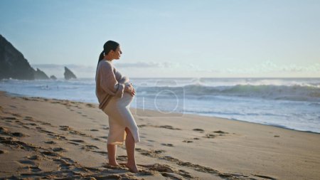 Schöne schwangere Frau posiert am Sandstrand und betrachtet die schäumenden Meereswellen am Sommerabend. Zarte werdende Mutter, die am schönen Meeresufer steht und ihren dicken Bauch streichelt. Glückliches Schwangerschaftskonzept.