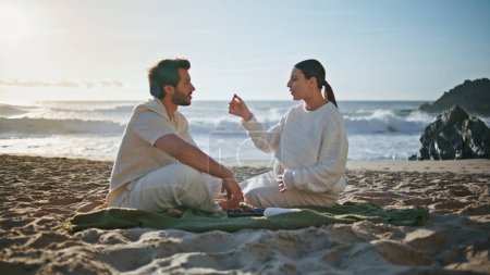 Spouses expecting baby enjoying calm evening picnic on sand sea coast. Loving pregnant wife feeding handsome husband sitting blanket together. Carefree happy couple touching belly relaxing on beach.