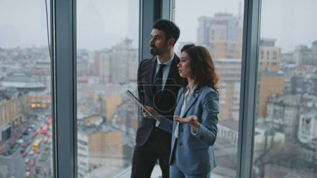Business partners speaking standing at office window looking on city view. Confident successful businesspeople discussing work project. Smart woman holding tablet with work plan talking to bearded man