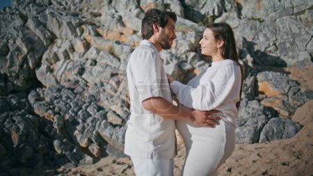 Pregnant pair enjoy nature weekend standing together at sunny seashore. Tender future parents hugging posing on summer rocky coast. Caring man embracing beloved woman expecting baby feeling affection.