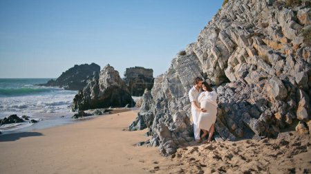 Gentle pregnant couple posing on rocky beach leaning at rough stones hugging. Happy carefree spouses expecting baby spending romantic weekend on beautiful seashore. Future parents enjoying pregnancy.