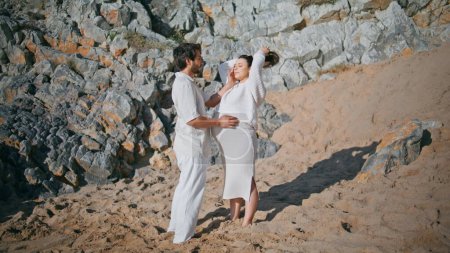 Family expecting baby standing on rocky coast enjoying sunny day together. Happy husband touching pregnant woman belly with tenderness. Lovely future mother hugging man relaxing on nature weekend.