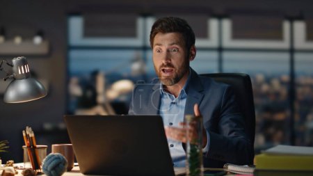Excited businessman explaining at online meeting with clients sitting dark office closeup. Successful emotional man concluding contract by video call. Elegant boss gesturing hands working at desk