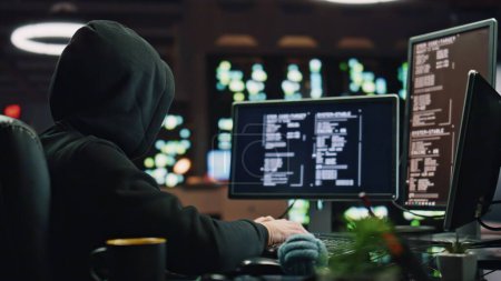 Anonymous hacker breaking cybersecurity at dark room closeup. Script kiddie in hoodie using algorithm source code looking monitors. Masked cybercriminal watching pc display reading data at night place