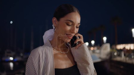 Joyful girl finish call waiting meeting at night city close up. Smiling stylish woman hung up smartphone standing at dark street alone. Happy brunette enjoy friendly conversation by cellphone outdoors