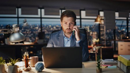 Confident boss calling mobile phone at evening city view office closeup. Focused man talking smartphone listening interlocutor at panoramic windows workplace. Serious businessman working late at night