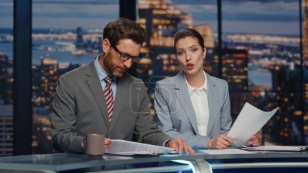 Couple reporters announcing breaking news in late multimedia channel closeup. Woman presenter broadcasting evening world events. Bearded anchorman drinking coffee. Smiling hosts discussing information