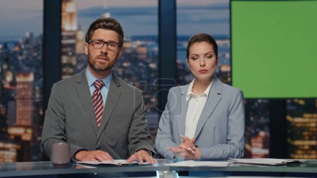 Photo for Serious news readers broadcasting chroma key tv newscast in night studio closeup. Positive charismatic presenters communicating with audience ending daily reportage. Anchorwoman winking to camera - Royalty Free Image
