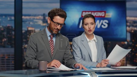 Happy anchors broadcasting news in evening studio closeup. Couple newsreaders reporting together lighting daily information at television channel. Bearded anchorman drinking tea anchorwoman talking