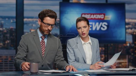 Confident hosts presenting daily breaking news in evening television show closeup. Serious professional presenters broadcasting newscast. Elegant man drinking tea woman reporting about business event