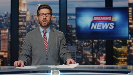 Photo for Smiling newscaster reporting business information in air in newsroom studio. Bearded anchor man talking at evening breaking news program. Anchorman holding script papers at television cable channel - Royalty Free Image