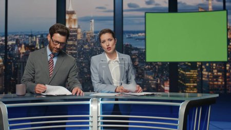 Couple newsreaders reporting green screen news in late multimedia channel closeup. Smiling woman presenter broadcasting current world events at chroma key studio. Bearded anchorman making coffee sip