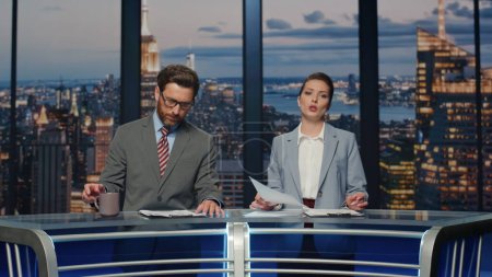 Couple hosts reporting breaking news in late multimedia channel closeup. Lady presenter broadcasting current world events. Bearded anchorman drinking coffee. Smiling reporters discussing information