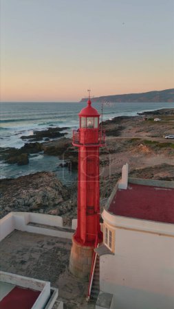 Aerial view beautiful beacon standing on stony seaside at summer twilight. High red lighthouse placed on scenic coastline for ships navigation. Picturesque pink sky over ocean waves vertically video.