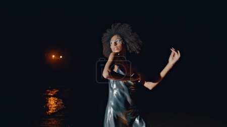 Sensuelle fille dansant nuit en robe de cocktail. Séduisante caméra afro-américaine jouissant d'un rythme sur la plage. Belle femme sexy performant dans la lumière. Magnifique modèle de fête en tenue argentée tendance