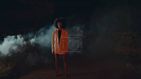 Modische Dame modelliert Nebelschwaden am Strand. Attraktive schlanke Frau mit Kamera im Scheinwerferlicht. Schlanke afrikanisch-amerikanische Sommernacht auf einer Outdoor-Festival-Party. Fabelhaftes Mädchen in Jacke