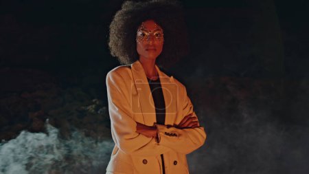 Glamouröse Frau modelliert Rauch mit glänzendem Make-up in der Nacht. Sexy Dame mit schwarzem Haar, Kamera mit verschränkten Händen. Stilvoller Afroamerikaner steht in Rauchwolke und posiert bei Outdoor-Festival-Licht.