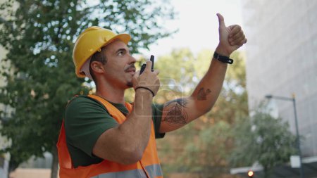 Photo for Uniform engineer showing thumb up talking walkie-talkie at urban district closeup. Confident foreman dictating messages to subordinates at modern exterior. Hardhat man working on architectural project - Royalty Free Image