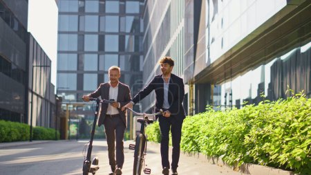 Two businessmen going city downtown. Smiling colleagues talking holding scooter bicycle at modern office center. Happy relaxed managers commuting home leaving shot. Corporate people lifestyle concept.