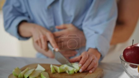 Affectionate couple preparing food at home kitchen close up. Beautiful woman slicing fresh apple for fruit salad. Tender man hugging happy wife cooking healthy breakfast. Young family weekend morning.