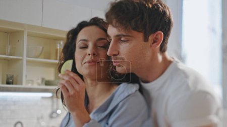 Spouses enjoy cooking together standing at modern comfortable kitchen close up. Smiling romantic couple tasting sweet apple slices on breakfast. Happy young man hugging beautiful wife weekend morning.