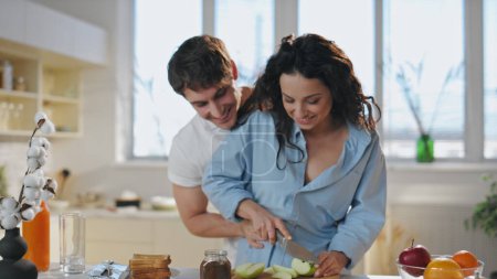Zärtliches Paar beim Kochen in der Küche, das sich lachend aus nächster Nähe umarmt. Liebevolle junge Ehepartner, die Spaß daran haben, morgens ein gesundes Frühstück zuzubereiten. Lächelnde Ehefrau schneidet mit Hilfe ihres Mannes frischen Apfel. 