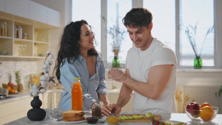 Wife tasting breakfast with husband leaning on kitchen countertop close up. Happy hugging couple eating apple slices together at home. Romantic smiling spouses enjoy healthy food at family weekend.